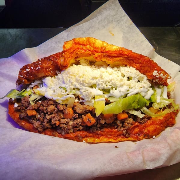 A large, vibrant Mexican gordita filled with ground meat, lettuce, and crumbled white cheese.